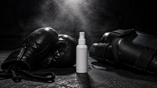 Hypochlorous acid cleaning spray bottle between boxing gloves and shin guards on a gym floor, mist in the air